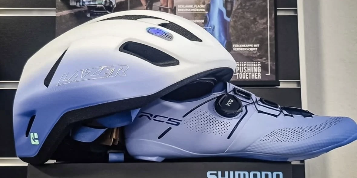 Der Lazer Sphere Helm in Weiß und Violett mit dem passenden Shimano RC5 Rennradschuh vor der Shimano-Wand im Laden